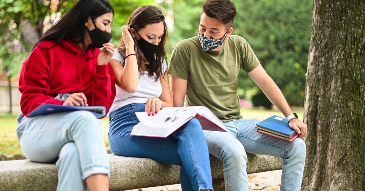Three students studying together sitting on a bench outdoor and wearing masks during coronavirus times